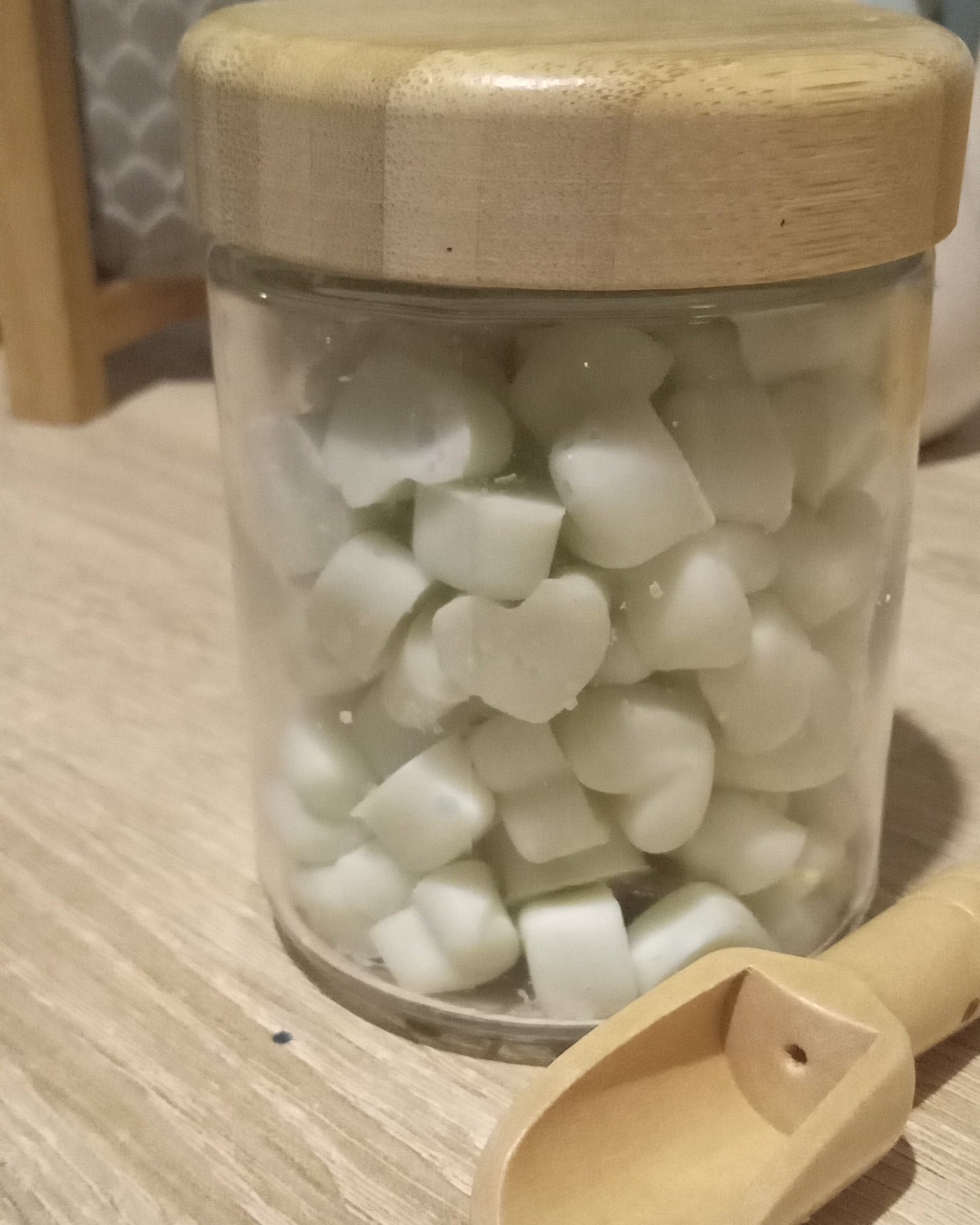 Jar of light green heart shaped mini wax melts with a wooden lid and scoop on a light wood surface