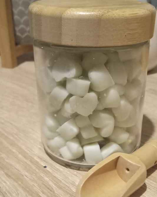 Jar of light green heart shaped mini wax melts with a wooden lid and scoop on a light wood surface