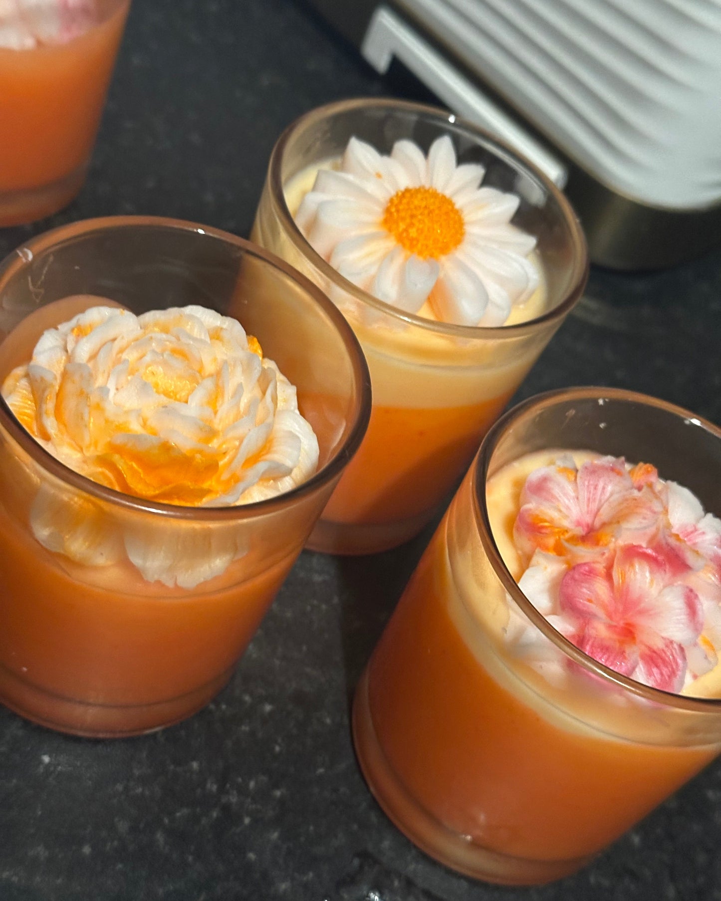 Three small glass containers of Tropical Delight scented coconut wax wickless candles with floral designs on a dark surface
